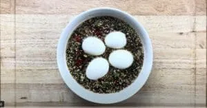 Boiled eggs in a savory soy sauce mixture with chopped vegetables and sesame seeds in a white bowl on a wooden table.